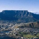 Aerial view of a coastal city where 10 Things to do in Cape Town come to life, with the prominent Table Mountain majestically in the background. The city boasts numerous waterfront buildings, green areas, and smaller hills. The ocean graces the foreground, completing this vibrant tableau.
