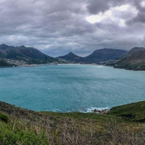 Panoramic view of a coastal bay with turquoise water surrounded by mountains under a cloudy sky. A small town is nestled between the mountains and the bay, offering a private cape peninsula tour experience, with lush greenery and a winding road in the foreground.