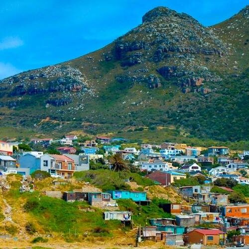 Colorful houses are scattered across a hillside, with lush green mountains in the background under a clear blue sky. Embark on a private Cape Peninsula tour to experience this vibrant community that seamlessly blends with the natural landscape, creating a picturesque scene.