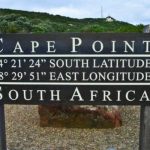 A sign at Cape Point, South Africa, marks coordinates 34° 21' 24" S and 18° 29' 51" E, a highlight of the Best of the Cape Tour. The lush greenery and cloudy sky in the background set a breathtaking scene.