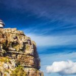 Cape of Good Hope A lighthouse perched atop a rugged cliff under a vivid blue sky with wisps of cloud, much like the scenic views on a Cape Peninsula tour. The landscape is dotted with sparse greenery, conveying solitude and natural beauty.