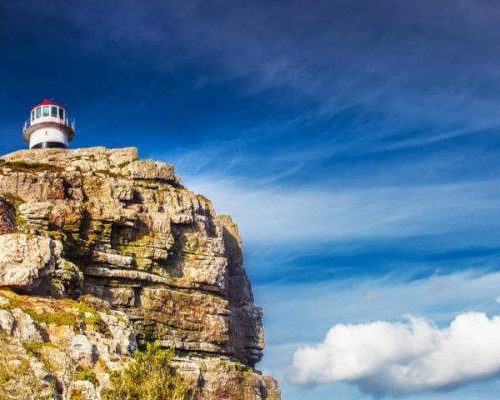 Cape of Good Hope A lighthouse perched atop a rugged cliff under a vivid blue sky with wisps of cloud, much like the scenic views on a Cape Peninsula tour. The landscape is dotted with sparse greenery, conveying solitude and natural beauty.