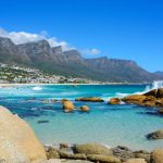 camps bay Experience the Best of the Cape Tour along a rocky shoreline with turquoise waters where waves crash against boulders. Rugged mountains rise in the distance under a clear blue sky, while sunlit, white buildings pepper the vibrant hillsides.