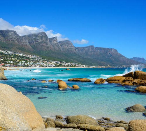 camps bay Experience the Best of the Cape Tour along a rocky shoreline with turquoise waters where waves crash against boulders. Rugged mountains rise in the distance under a clear blue sky, while sunlit, white buildings pepper the vibrant hillsides.