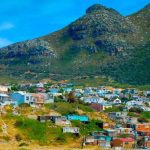 HOUT BAY Colorful houses are scattered across a hillside, with lush green mountains in the background under a clear blue sky. Embark on a private Cape Peninsula tour to experience this vibrant community that seamlessly blends with the natural landscape, creating a picturesque scene.