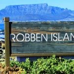 A sign reading Robben Island stands amid rocky terrain and green plants, offering a glimpse into the iconic Robben Island and Table Mountain tour. In the background, a body of water and majestic mountain range stretch beneath a clear blue sky.
