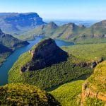 Explore a breathtaking view of a lush green valley with a winding river, surrounded by steep, verdant mountains under a clear blue sky. This travel guide to South Africa highlights landscapes dominated by a prominent peak and serene, expansive horizons.