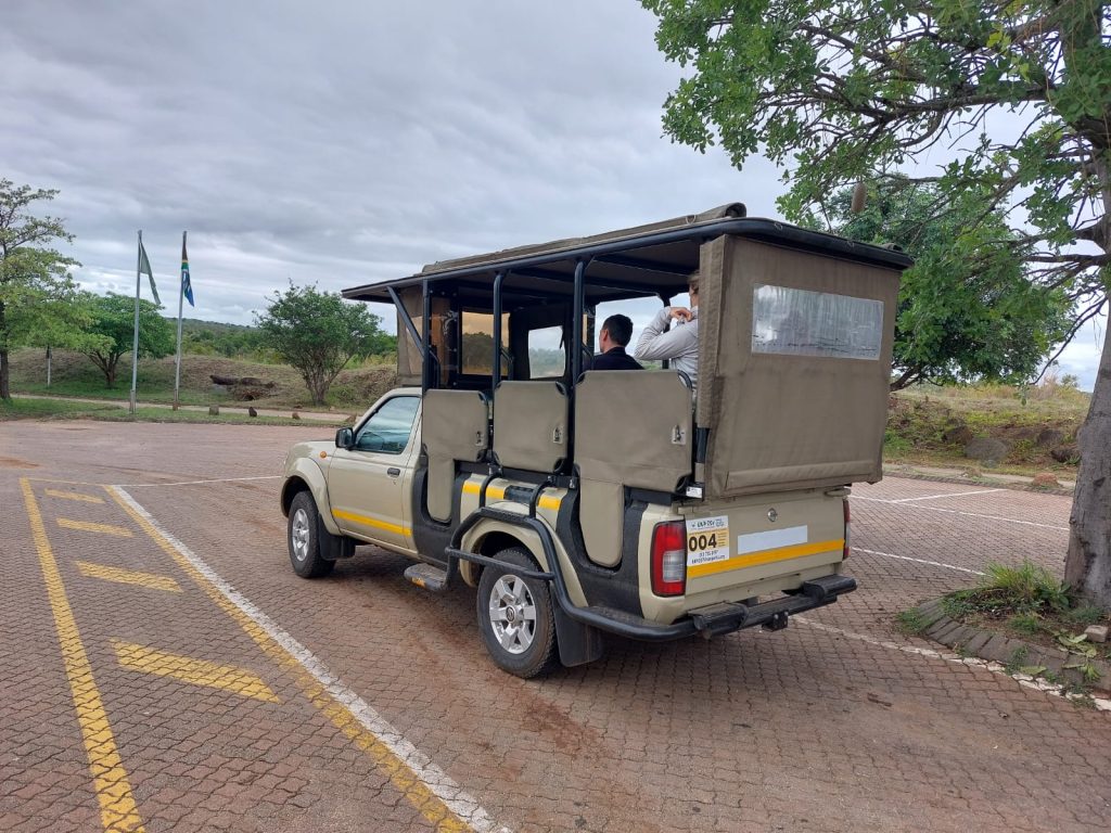 A safari vehicle is parked, ready for a tour in Kruger National Park. It has open sides and a canvas roof, with passengers seated inside. The ground is paved with a few trees and partly cloudy skies in the background. Flags are visible in the distance.