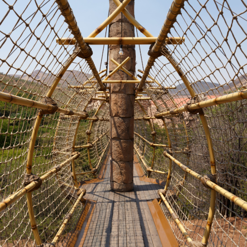 A narrow bridge made of wooden planks and ropes stretches across a canopy of green trees. The ropes form a protective netting on both sides. Distant mountains are visible under a clear blue sky.