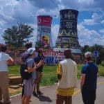A group of people stand on a path observing colorful painted towers against a cloudy sky. One tower features a vibrantly decorated advertisement, while the other depicts the words Soweto with various graphics. Trees and a road are in the background.