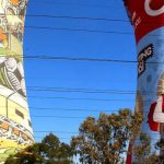 Colorful murals cover two large cooling towers in Soweto. One mural features musicians and dancers, while the other showcases people reading with the phrase Empowering Education. Trees and power lines are in the foreground against a clear blue sky, celebrating the vibrant spirit of Soweto.