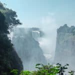 A majestic waterfall cascades between rocky cliffs, surrounded by lush green foliage. Mist rises from the base, creating a dreamy atmosphere under a clear blue sky.