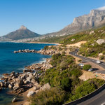 A winding coastal road hugs the shoreline, with cars driving along. The landscape features rocky beaches, blue ocean waters, and rugged mountains under a clear blue sky. Lush greenery lines the road, with a distinctive mountain peak in the background.