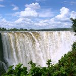 A powerful waterfall cascades over a wide cliff under a bright blue sky with scattered clouds. Lush green foliage surrounds the foreground, framing the view of the majestic waterfall.