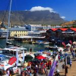 A lively waterfront bustling with tourists exploring Cape Town tour packages, boats docked in a harbor, and a large Ferris wheel. The scene is set against majestic mountains under a clear blue sky, with vibrant shops and people enjoying the day.