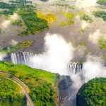 Aerial view of a large waterfall surrounded by lush greenery and mist. The river flows over a wide cliff, creating a striking cascade. A road with a bridge curves through the foreground, while clouds drift above.