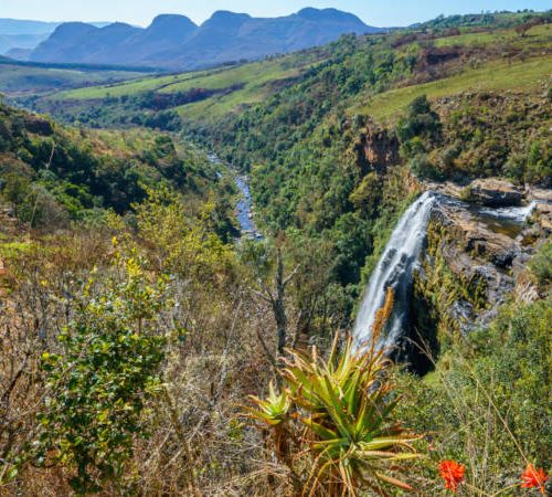 A breathtaking view of the Berlin Falls in South Africa, cascading down a rugged cliff amidst lush greenery. The river winds through a verdant valley, with mountain peaks visible in the distance under a clear blue sky.