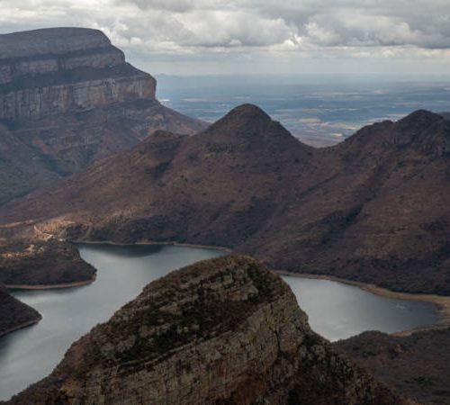 Aerial view of a scenic landscape with rugged mountain ranges and a winding river below. The sky is overcast, casting soft, diffused light over the terrain, highlighting the textures and contours of the rocky formations.