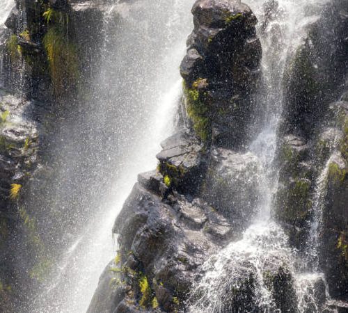 A cascading waterfall flows over dark, jagged rocks, creating mist in its descent. Sunlight highlights patches of green moss on the rocks, adding a touch of color to the otherwise rocky landscape.