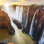 Majestic waterfall cascading over a series of jagged cliffs with lush greenery, creating misty spray and a river below. The scene is vibrant with warm hues, capturing the powerful and serene beauty of nature at sunset.