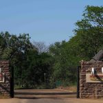 A rustic entrance with stone pillars and signs featuring tusks on either side of an open road, surrounded by lush green trees under a clear blue sky. An A-frame building is visible on the right side.
