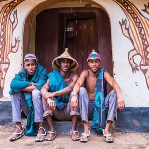 Three men in traditional hats and colorful fabrics sit on a step before a decorated building at Lesedi Cultural Village. The intricate animal designs on the wall frame their relaxed presence, vividly showcasing cultural attire and heritage amidst this vibrant community hub.